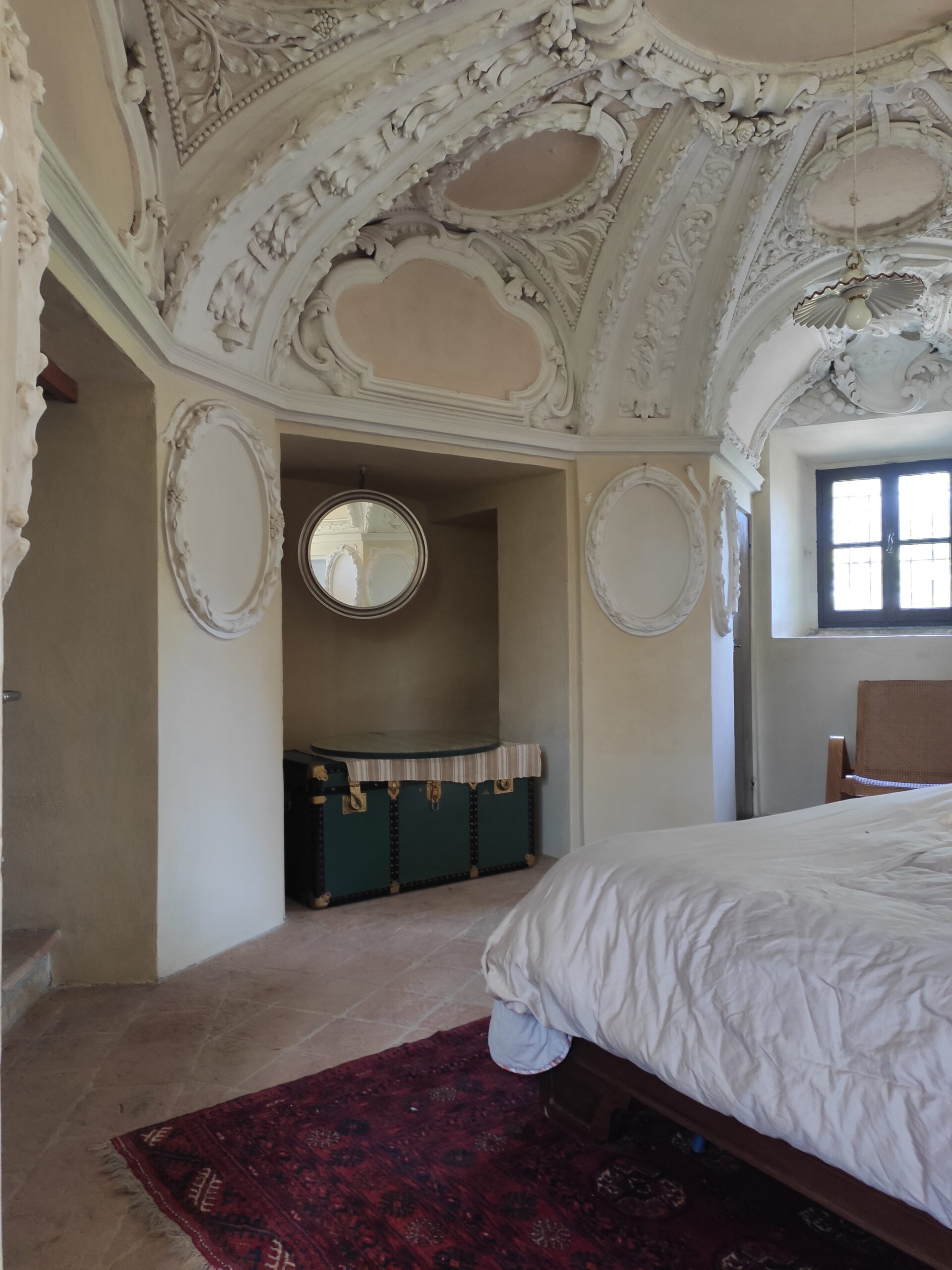 Room with a window showing the corner of the bed, a round mirror and a red carpet.